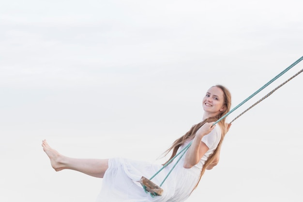 Young happy woman in white dress riding a swing on sky background