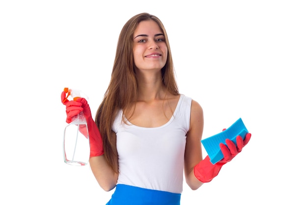 Young happy woman in white shirt and blue apron with red gloves holding duster and detergent on white background in studio