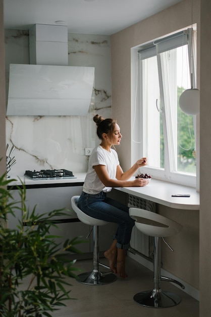 Young happy woman in a white Tshirt and jeans holds a plate of cherries in her hands and takes a berry at home in the kitchen