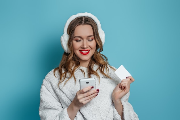 Young happy woman in winter white faux fur coat and fur headphones holds mobile phone and credit card and makes online order, shopping 