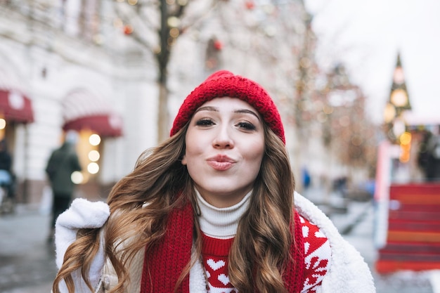 Young happy woman with curly hair in red knitted hat at the Christmas fair in winter street decorated with lights