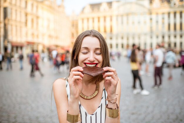 Photo young and happy woman with dark chocolate bar standing outdoors on the grand place in brussels in belgium. belgium is famous of its chocolate