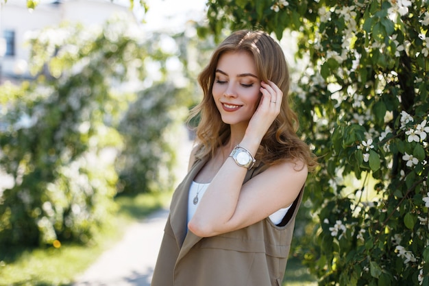 Young happy woman with a wonderful smile in a stylish beige vest in a white T-shirt posing outdoors with blooming green trees. Beautiful girl enjoy nature in summer day