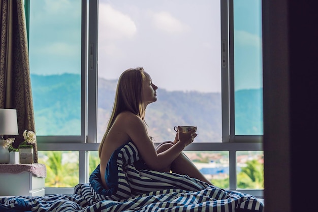 Young happy woman woke up in the morning in the bedroom by the window with beautiful mountain views.