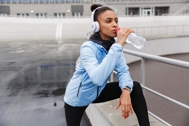 young healthy african sportswoman listening to music with wireless earphones wrking out at the stadium, holding water bottle