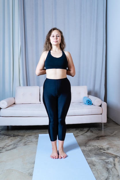 Young healthy beautiful woman in sports top and leggings practicing yoga at home standing in pose on yoga mat