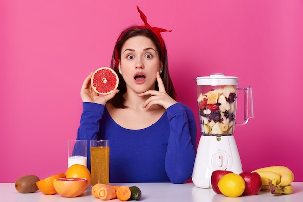 Young and healthy woman has surprising facial expression, holds piece of grapefruit in her hand, isolated on pink. Large variety of fresh fruit and vegetables on table surface.