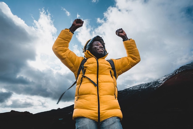 A young hiker man of African ethnicity in a yellow vest and backpack rejoices with his arms raised concept of victory wanderlust and achievement