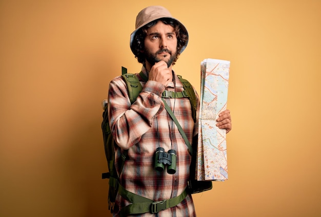 Young hiker man with curly hair and beard hiking wearing water canteen using city map serious face thinking about question very confused idea