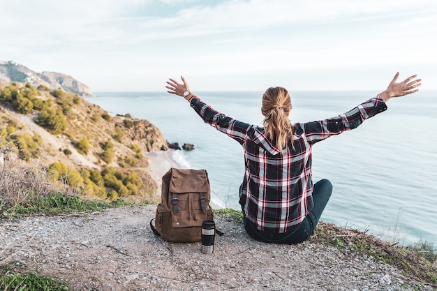 Young hip woman with open arms and a backpack explores the coast on a beautiful day. Concept of exploration and adventures