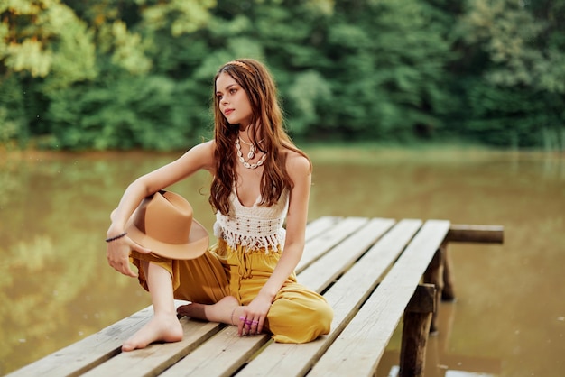 A young hippie woman sits on a lake bridge wearing stylish eco clothes and smiling