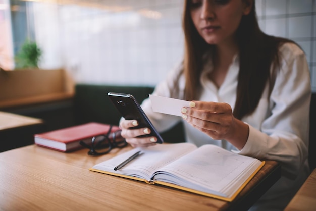 Young hipster girl 20s holding smartphone in hand and looking at business card for dialing