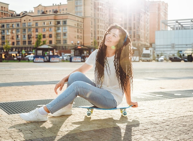 A young hipster girl wearing headphones sitting on a skateboard spends time in the city at sunset evening autumn sunset generation z