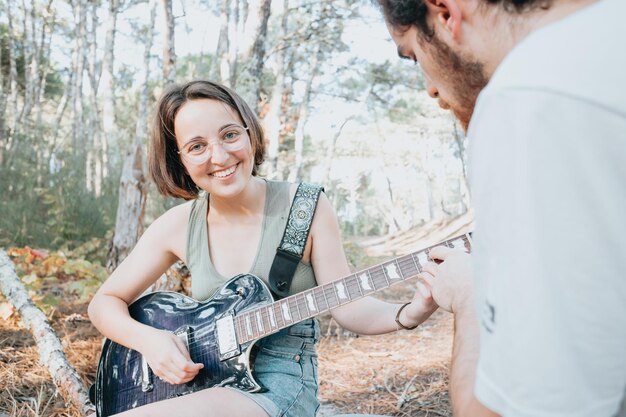 Young hipster woman learning to play the guitar smiling with boyfriend teacher outside park of the city. Having fun learning a new skill, music play seasonal style. Young short hair girl. Copy space