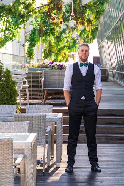 A young hipsterlooking man in a suit with a bow tie a manager in a summer street cafe waits for visitors before opening