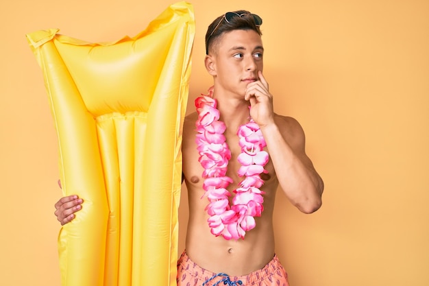 Young hispanic boy wearing swimsuit and holding summer matress float serious face thinking about question with hand on chin, thoughtful about confusing idea