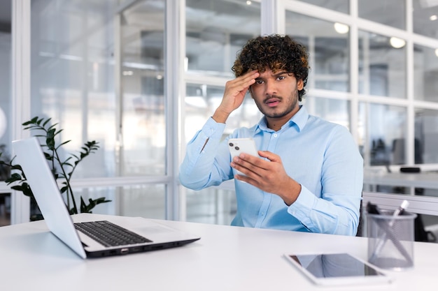 Young hispanic businessman sitting at desk in office holding phone worriedly holding head and