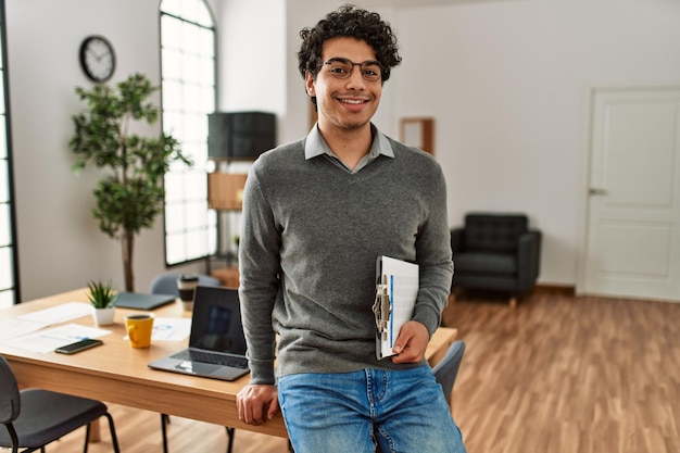 Young hispanic businessman smiling happy holding clipboard at the office.