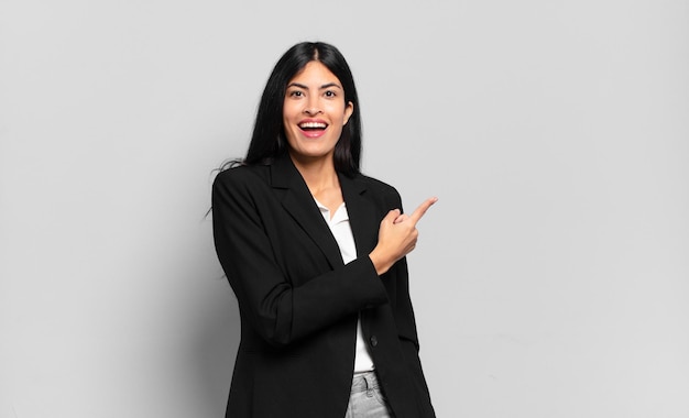 Young hispanic businesswoman looking excited and surprised pointing to the side and upwards to copy space