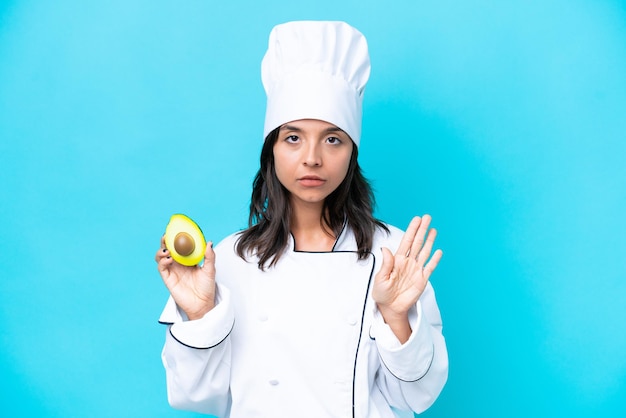 Young hispanic chef woman holding avocado isolated on blue background making stop gesture