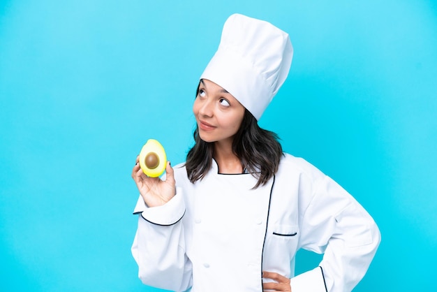 Young hispanic chef woman holding avocado isolated on blue background thinking an idea while looking up