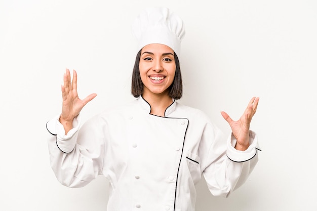 Young hispanic chef woman isolated on white background receiving a pleasant surprise excited and raising hands