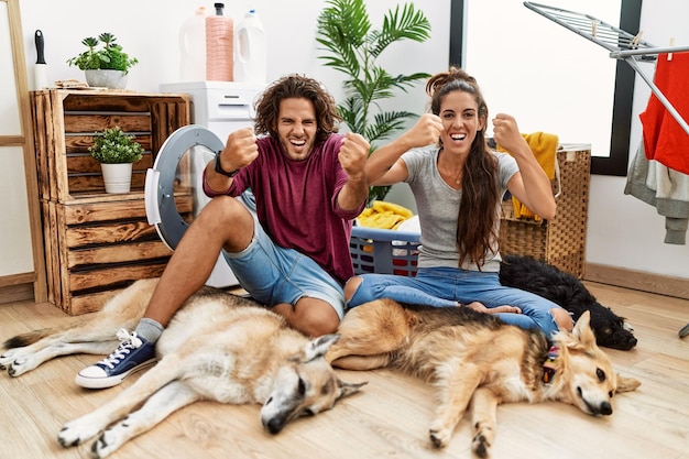 Young hispanic couple doing laundry with dogs angry and mad raising fists frustrated and furious while shouting with anger rage and aggressive concept