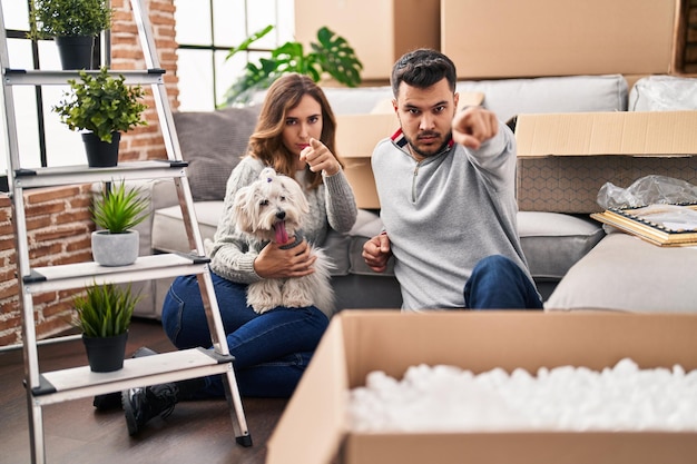 Young hispanic couple sitting on the floor at new home with log pointing with finger to the camera and to you confident gesture looking serious