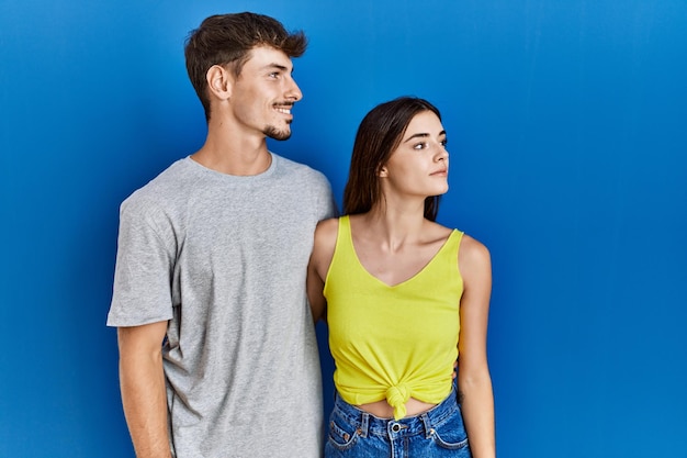 Young hispanic couple standing together over blue background looking to side, relax profile pose with natural face and confident smile.