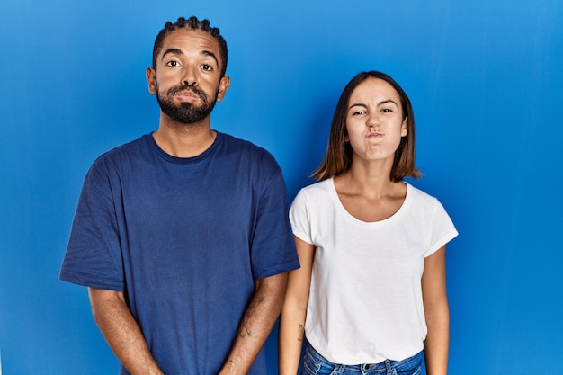 Young hispanic couple standing together puffing cheeks with funny face. mouth inflated with air, crazy expression.