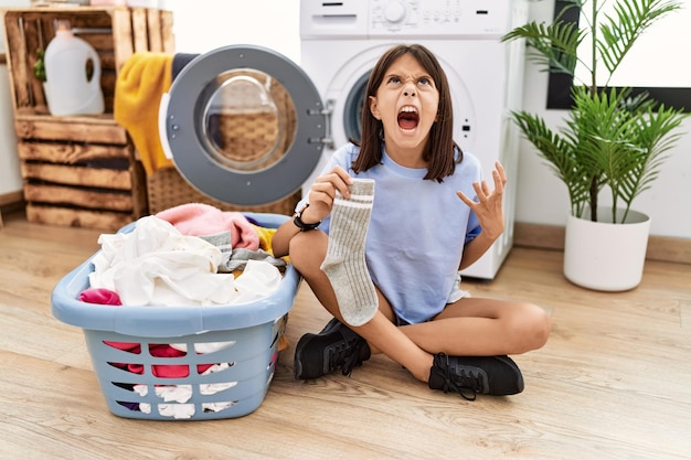 Photo young hispanic girl doing laundry holding socks crazy and mad shouting and yelling with aggressive expression and arms raised. frustration concept.