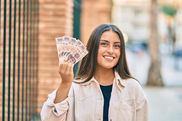 Young hispanic girl smiling happy holding mexican 500 pesos banknotes at the city