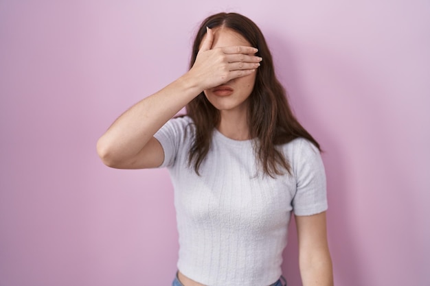 Young hispanic girl standing over pink background covering eyes with hand looking serious and sad sightless hiding and rejection concept