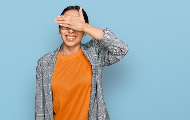 Young hispanic girl wearing business jacket and glasses smiling and laughing with hand on face covering eyes for surprise blind concept