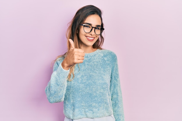 Young hispanic girl wearing casual clothes and glasses smiling happy and positive thumb up doing excellent and approval sign