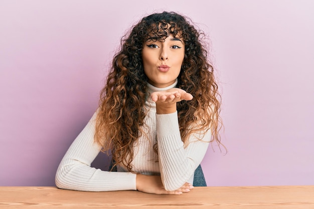 Young hispanic girl wearing casual clothes sitting on the table looking at the camera blowing a kiss with hand on air being lovely and sexy love expression