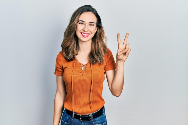 Young hispanic girl wearing casual clothes smiling with happy face winking at the camera doing victory sign number two