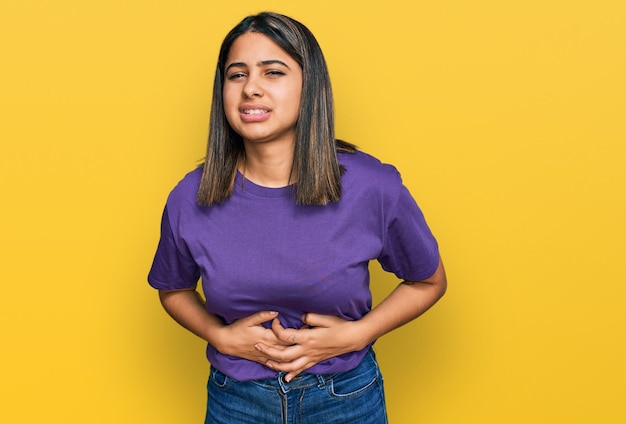 Young hispanic girl wearing casual purple t shirt with hand on stomach because nausea, painful disease feeling unwell. ache concept.