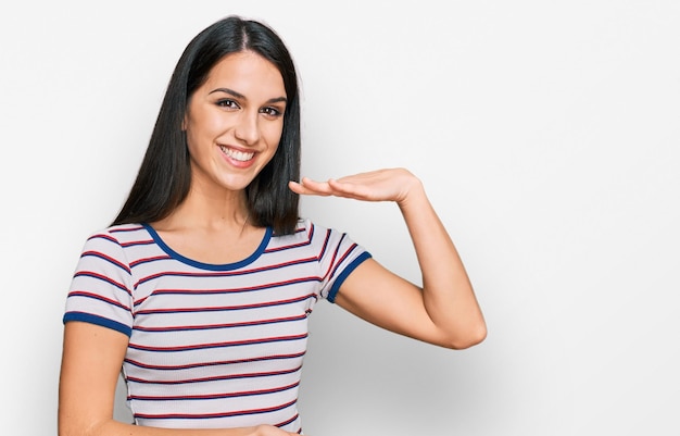 Young hispanic girl wearing casual striped t shirt gesturing with hands showing big and large size sign, measure symbol. smiling looking at the camera. measuring concept.