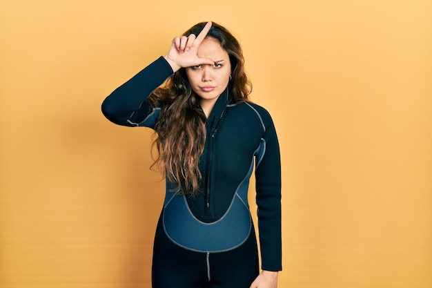 Young hispanic girl wearing diver neoprene uniform making fun of people with fingers on forehead doing loser gesture mocking and insulting.