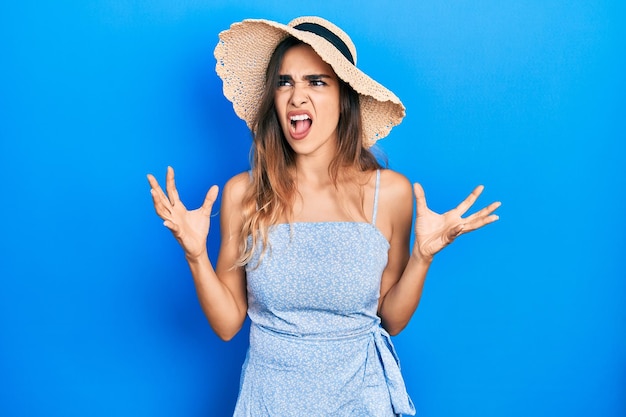 Young hispanic girl wearing summer hat crazy and mad shouting and yelling with aggressive expression and arms raised frustration concept