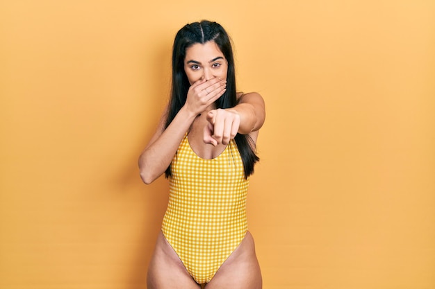 Young hispanic girl wearing swimsuit laughing at you pointing finger to the camera with hand over mouth shame expression