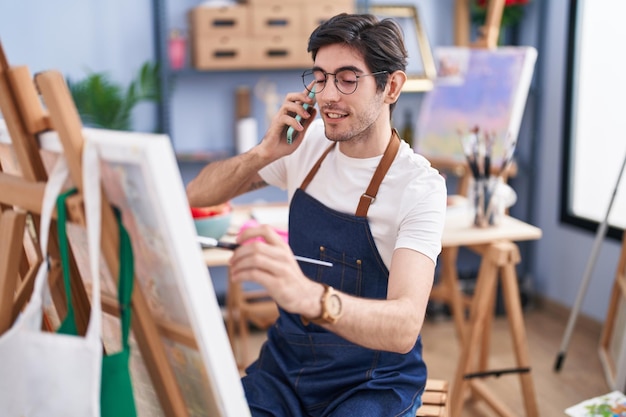 Young hispanic man artist talking on smartphone drawing at art studio