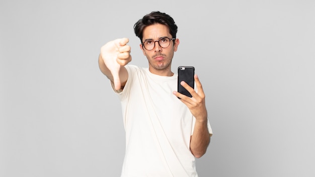 Young hispanic man feeling cross,showing thumbs down and holding a smartphone