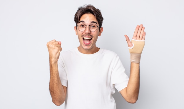 Young hispanic man feeling shocked,laughing and celebrating success. hand bandage concept