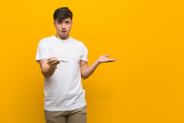 Young hispanic man holding an airplane icon impressed holding copy space on palm.