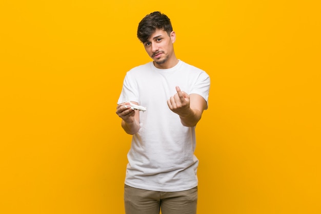 Young hispanic man holding an airplane icon pointing with finger at you as if inviting come closer.