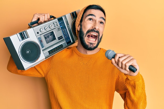 Young hispanic man holding boombox listening to music singing with microphone angry and mad screaming frustrated and furious shouting with anger looking up