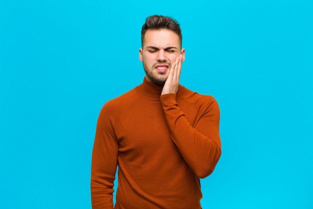 Young hispanic man holding cheek and suffering painful toothache, feeling ill, miserable and unhappy, looking for a dentist against blue wall