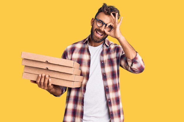 Young hispanic man holding delivery pizza box smiling happy doing ok sign with hand on eye looking through fingers